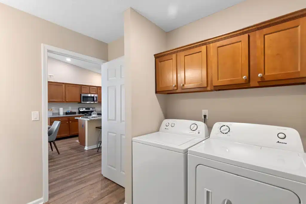 A laundry room with a washing machine and dryer below wooden cabinets, adjacent to a kitchen visible through an open door.