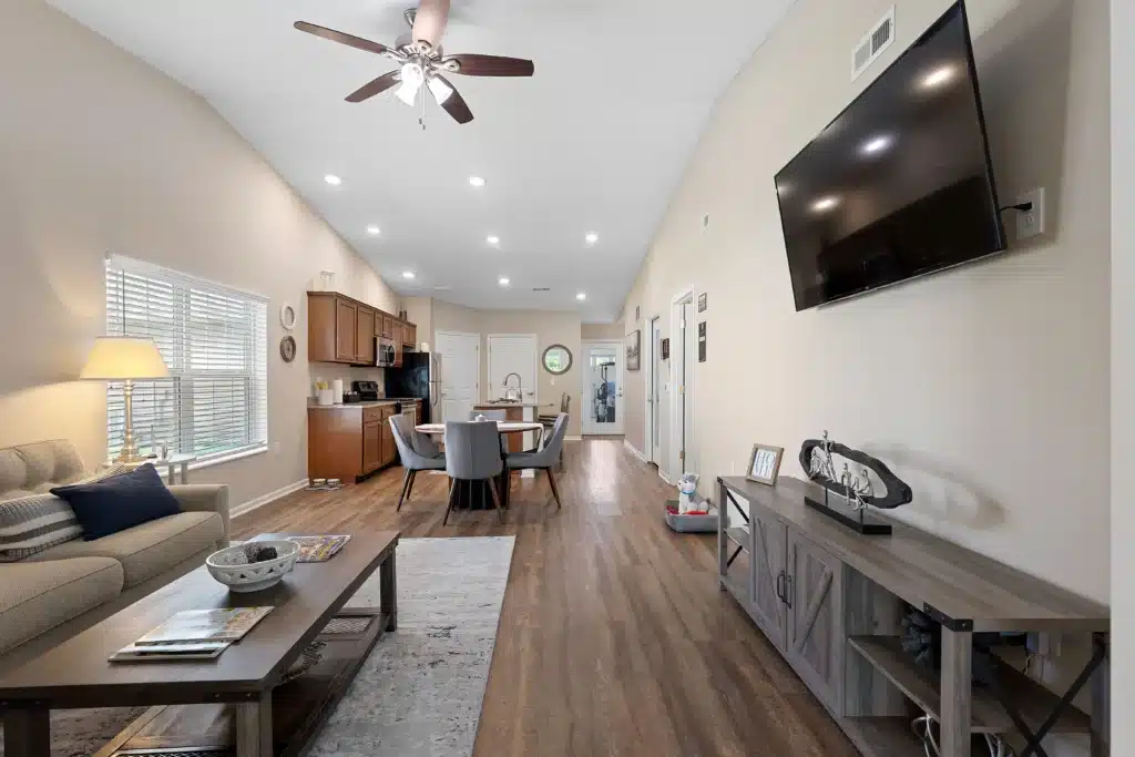 Open-concept living and dining area with wood flooring, beige walls, a mounted TV, kitchen in the background, and natural light from a window.