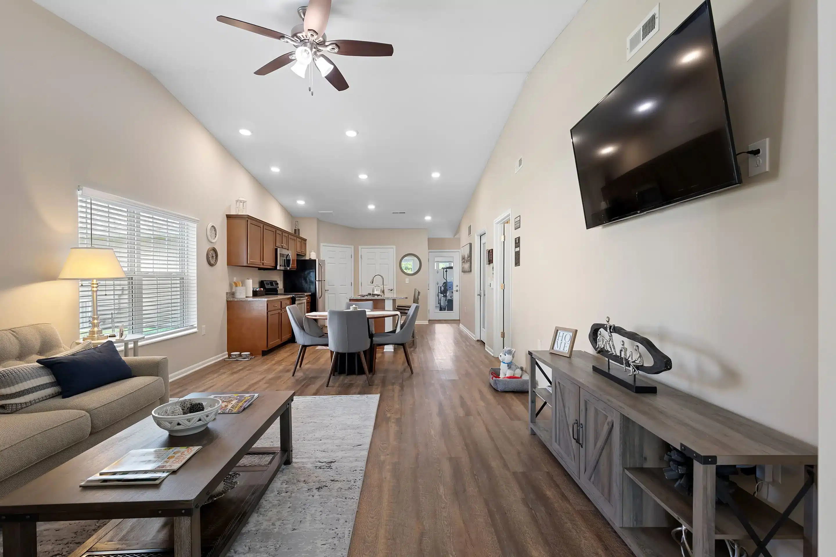Open-concept living and dining area with wood flooring, beige walls, a mounted TV, kitchen in the background, and natural light from a window.