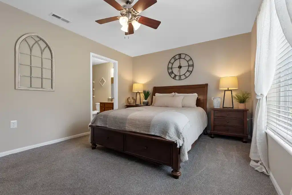 A neatly made bed with beige bedding sits between two nightstands with lamps in a carpeted bedroom with neutral walls and a ceiling fan. A window with sheer curtains lets in natural light.