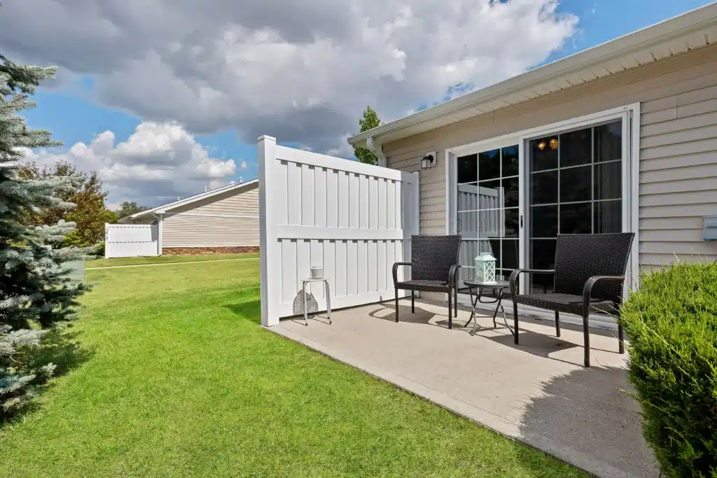 Small concrete patio with two black chairs, a side table, and a white privacy fence outside a beige house with sliding glass doors and a well-kept lawn.