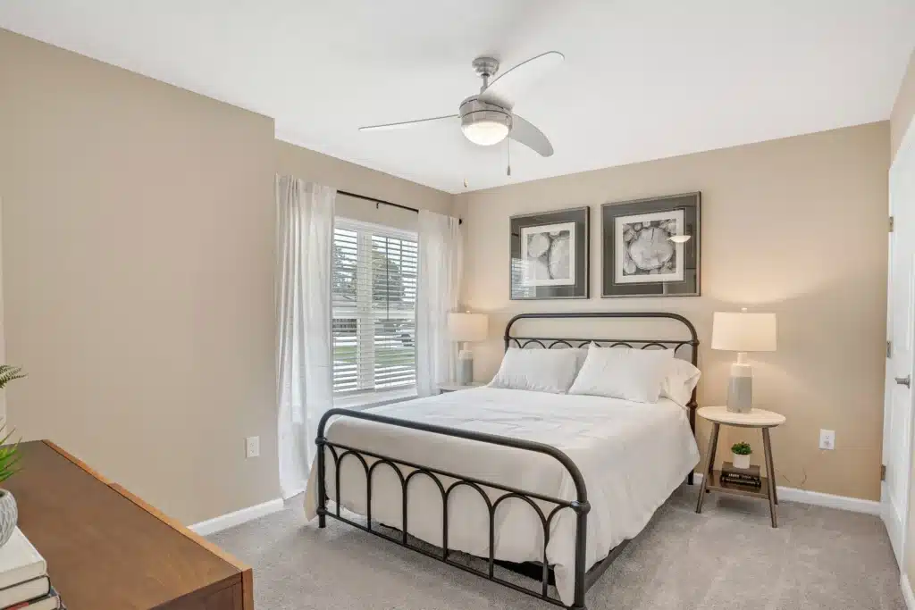 A neatly made bed with white bedding, two lamps on side tables, framed art above the headboard, and a ceiling fan in a beige-walled bedroom with a window.