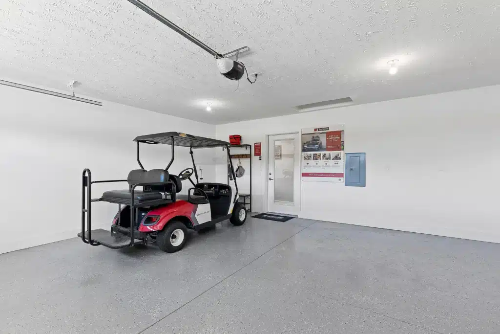 A red golf cart is parked inside a clean, spacious garage with gray floors, white walls, and overhead lighting. Posters and storage items are visible on the back wall.