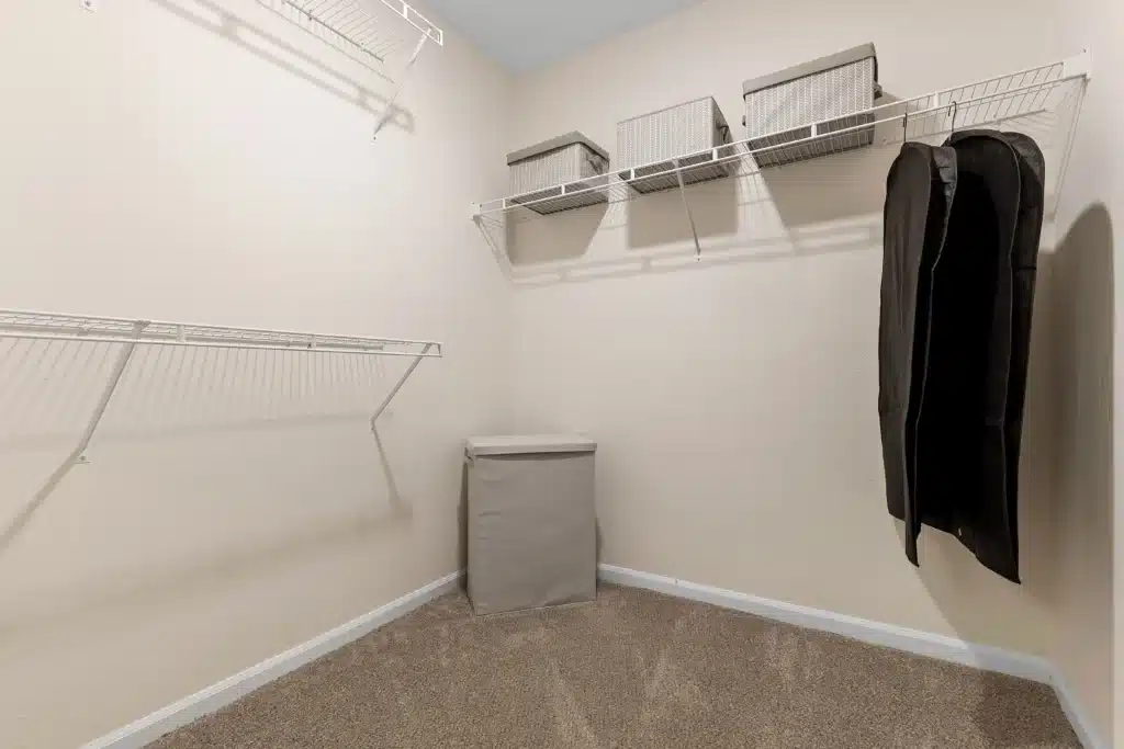 Empty walk-in closet with beige carpet, off-white walls, wire shelves holding gray storage bins, a hamper, and a black garment bag hanging on the right side.