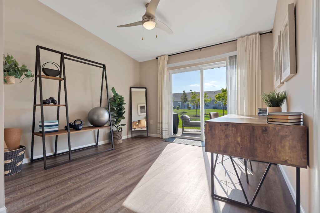 A modern, sunlit home office features a wooden desk, open shelving with decor, a large mirror, a potted plant, and sliding glass doors leading to an outdoor area.