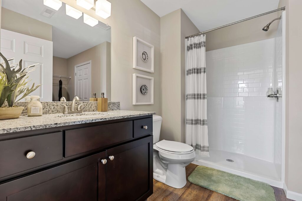 Modern bathroom with a granite countertop, dark wood vanity, toilet, shower with white tile and striped curtain, green rug, and wall art above the toilet.