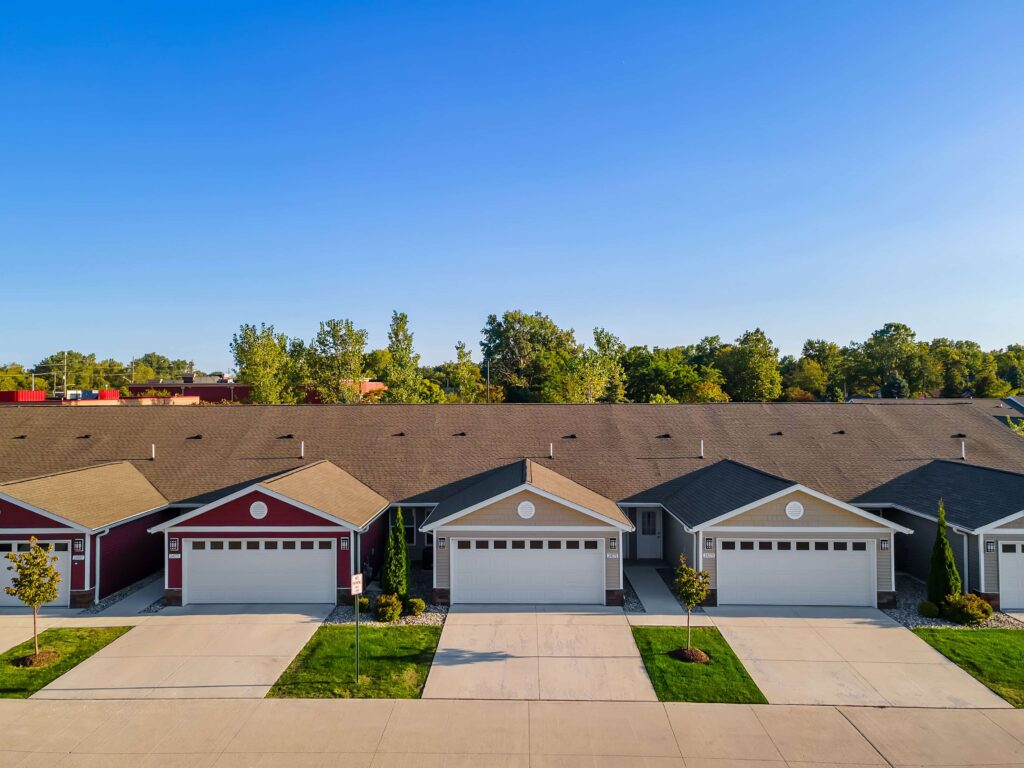 Row of attached single-story houses with garages and driveways, surrounded by trees, under a clear blue sky.