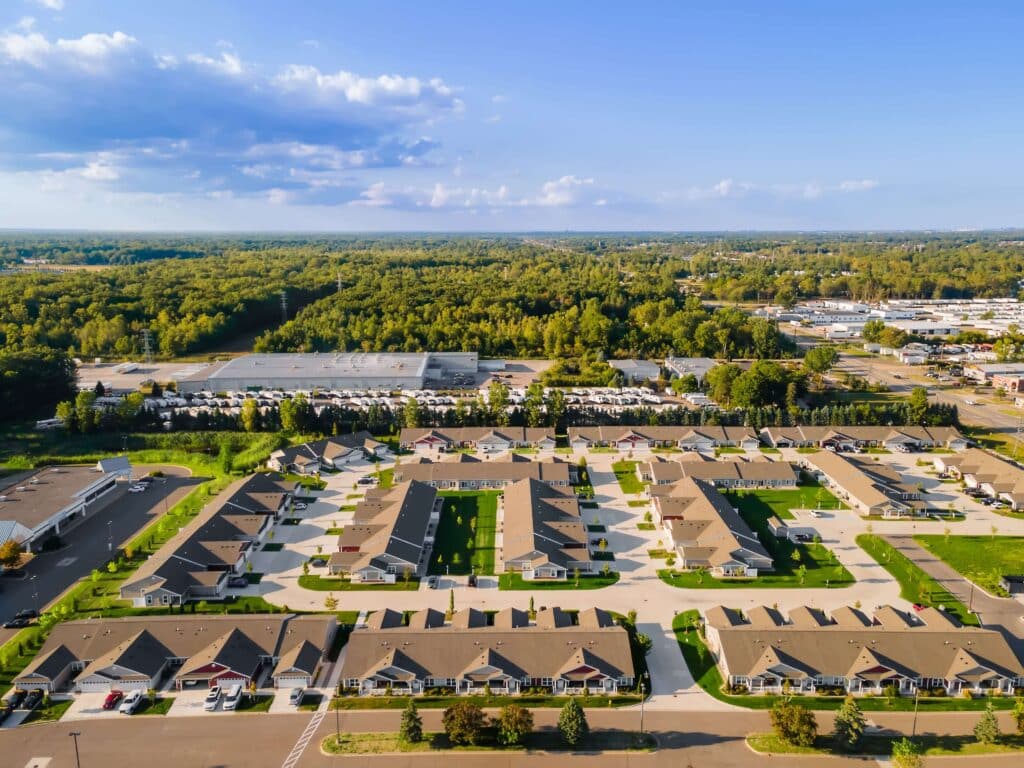 Aerial view of a suburban residential neighborhood with rows of single-story houses, surrounded by trees and nearby commercial buildings under a clear sky.