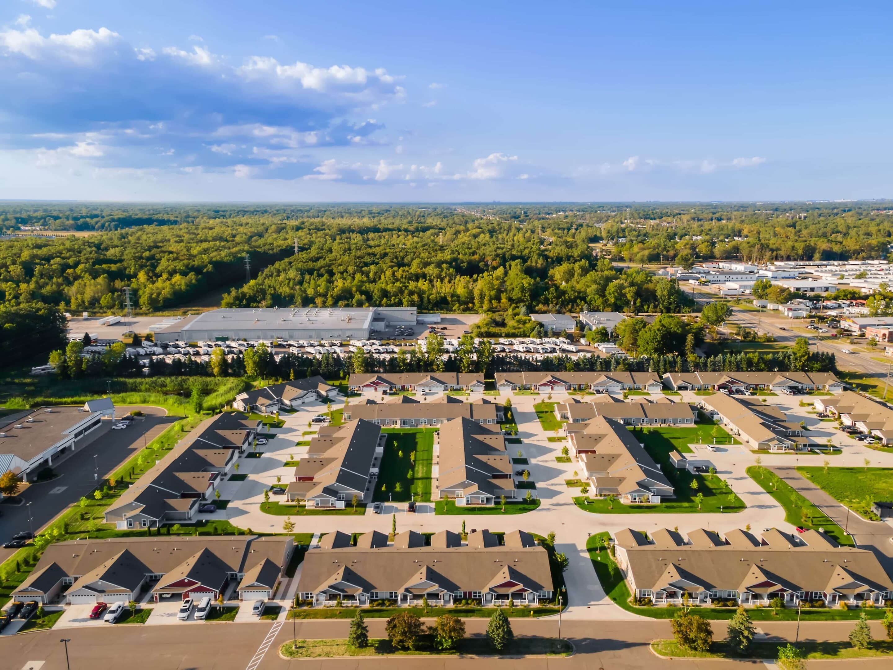 Aerial view of a suburban residential neighborhood with rows of single-story houses, surrounded by trees and nearby commercial buildings under a clear sky.