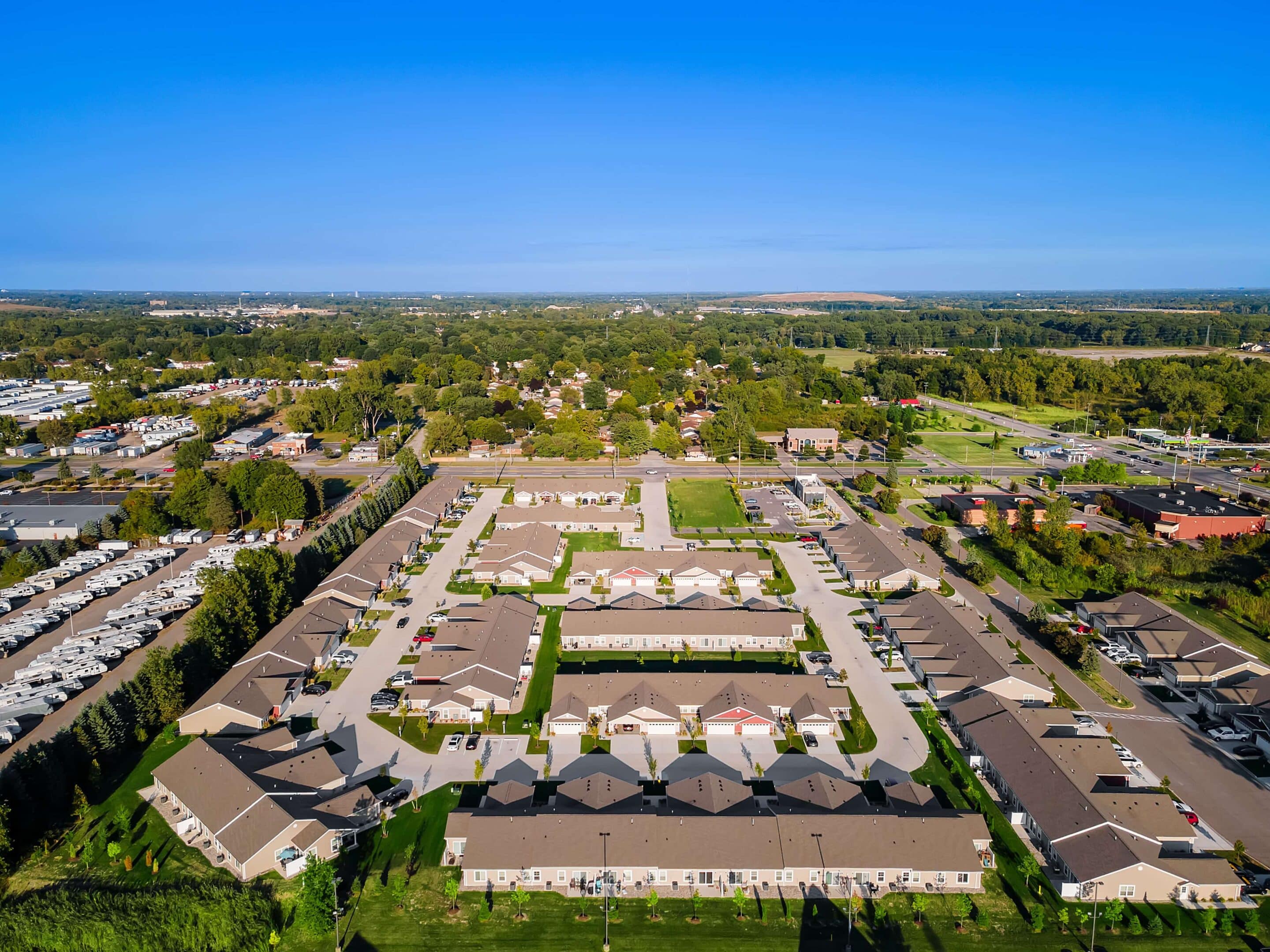 Aerial view of a suburban residential complex with multiple single-story buildings arranged around driveways and green lawns, surrounded by trees and other buildings.