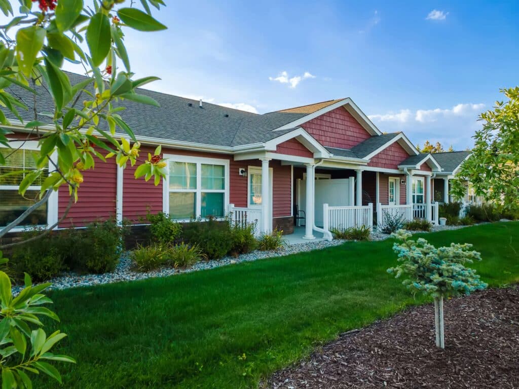 Single-story building with red siding, white trim, and a covered front porch set in a landscaped yard with green grass and small shrubs.