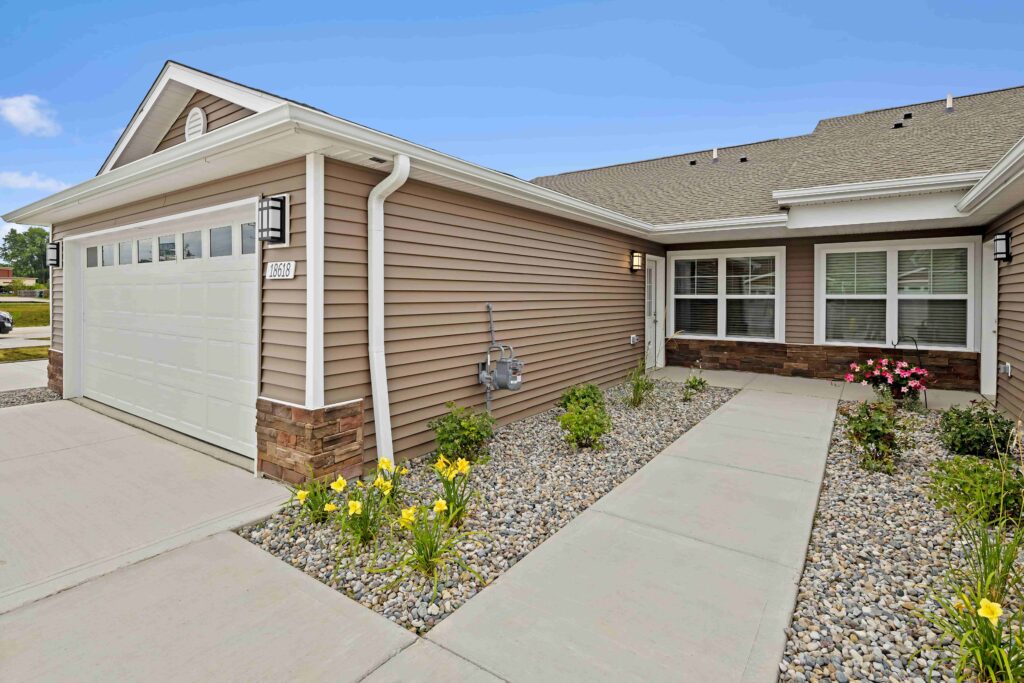 Single-story house with beige siding, a two-car garage, and a concrete walkway bordered by rocks and small plants leading to the front entrance.