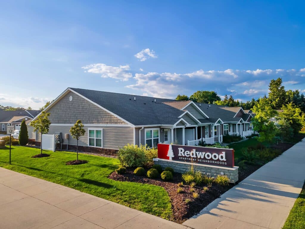 Single-story apartment buildings with gray siding and maintained landscaping; a sign in front reads "Redwood Apartment Neighborhoods" on a sunny day.