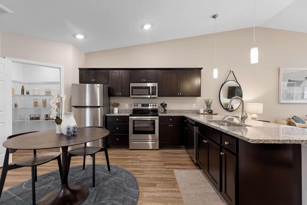 Modern kitchen with dark cabinets, stainless steel appliances, granite countertops, and a round dining table on a rug; pendant lights hang over the counter.