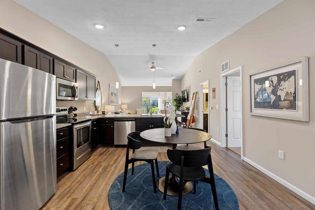 Modern kitchen and dining area with stainless steel appliances, dark cabinets, round table with four chairs, wood flooring, and neutral walls.