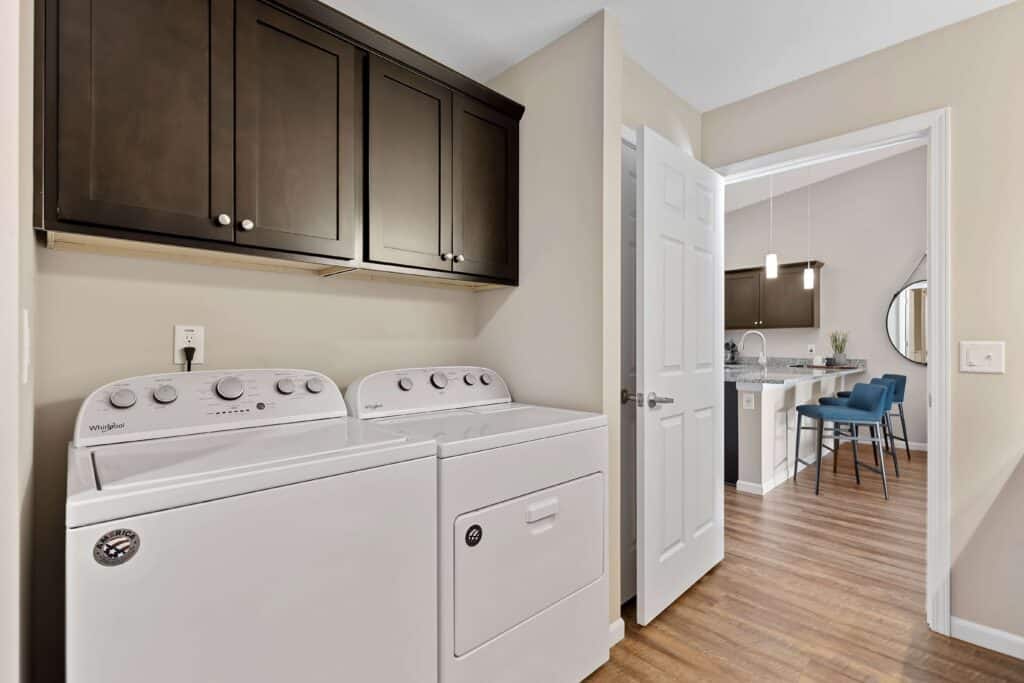Laundry area with a white washer and dryer below dark wood cabinets, adjacent to an open door leading to a modern kitchen and dining area with wood flooring.