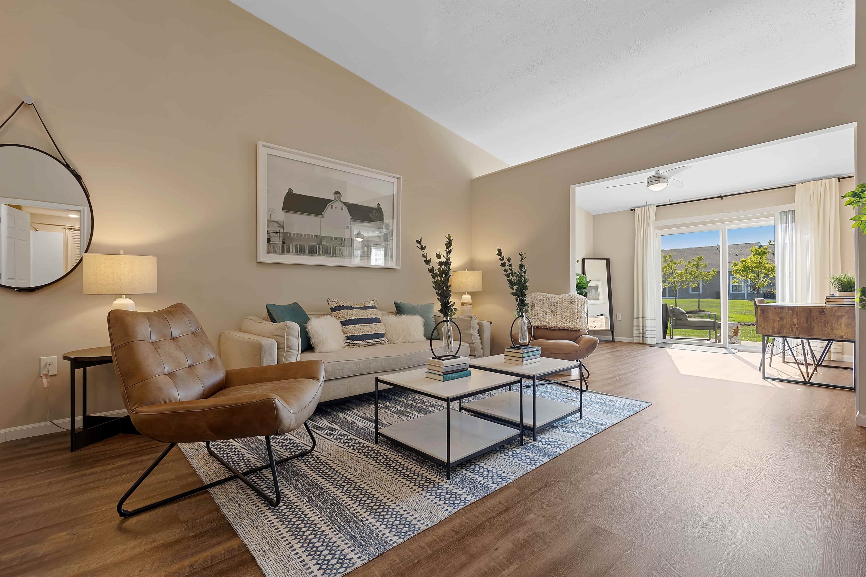 Modern living room with beige walls, brown leather chairs, a cream sofa, coffee tables, and large windows letting in natural light. A desk and chair are positioned near the sliding glass door.
