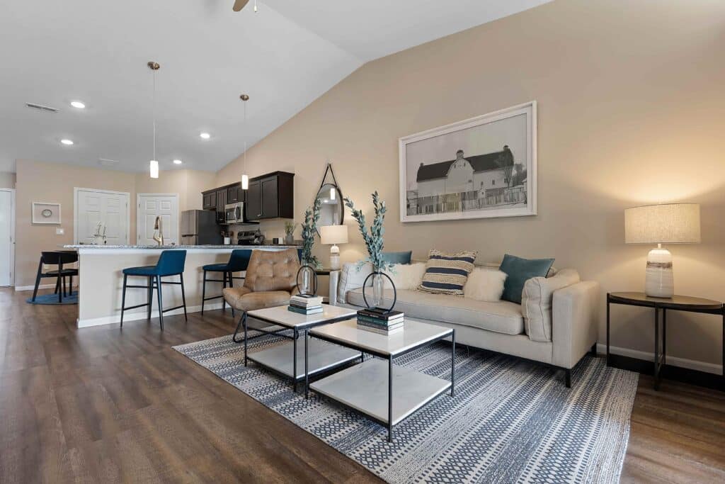 Modern living room with neutral decor, a beige sofa, armchair, coffee table, and rug; open kitchen with bar stools and pendant lights in the background.