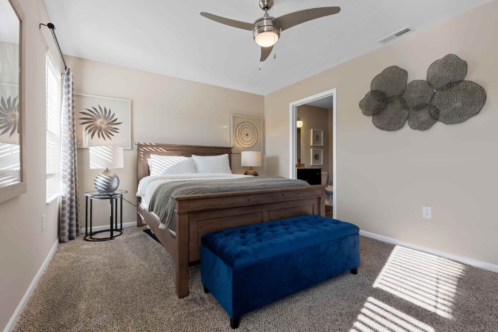 A bedroom with a wooden bed, white bedding, a blue tufted bench, beige carpet, modern wall art, a ceiling fan, and sunlight streaming through a window.