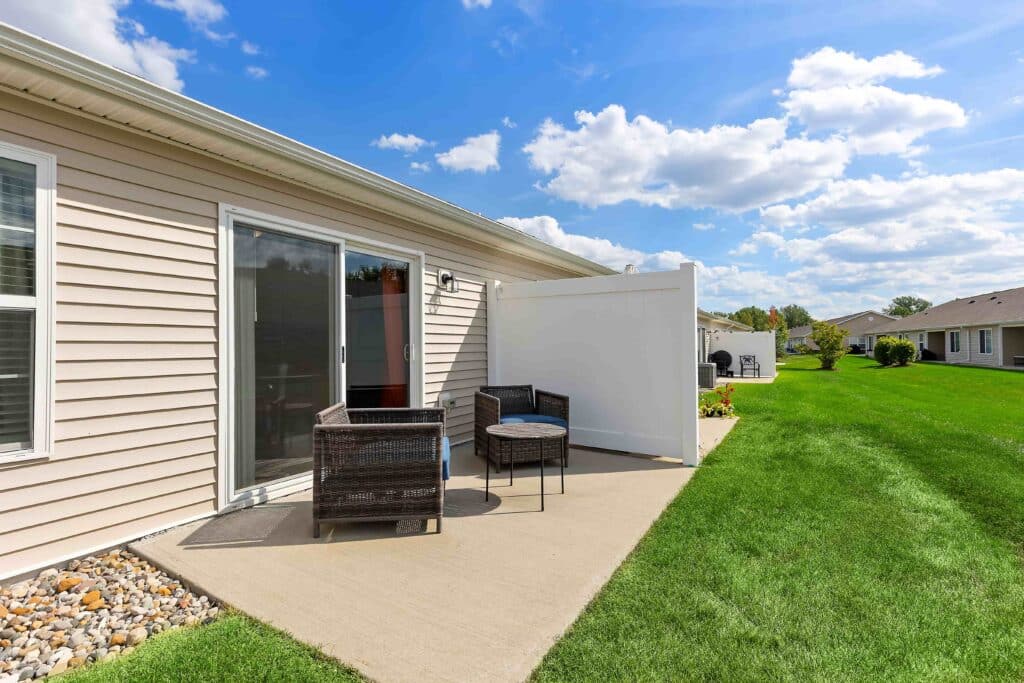 A concrete patio with two wicker chairs, a small table, and a privacy screen sits beside a beige house, with a green lawn and blue sky in the background.