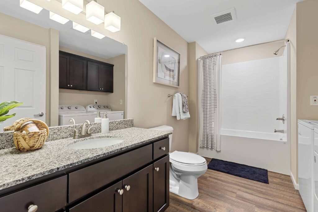 Modern bathroom with granite countertop, dark wood cabinets, toilet, and shower-tub combo; laundry machines are visible in an adjacent room.