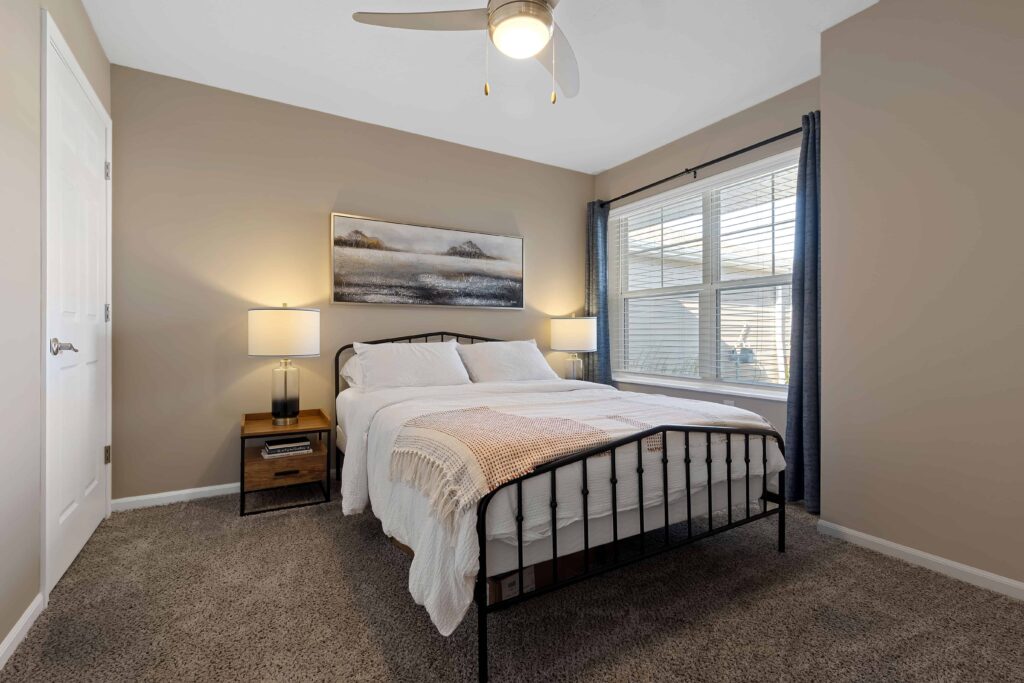 A neatly made bed with white bedding and a metal frame is centered in a carpeted bedroom with beige walls, two nightstands, lamps, a window with curtains, and a ceiling fan.