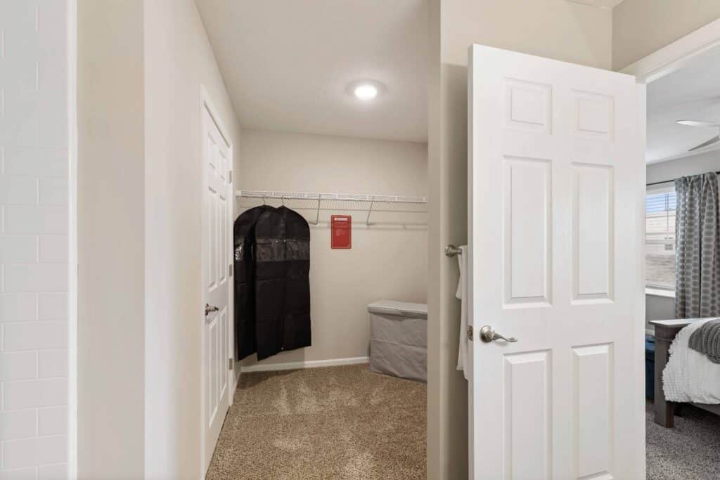 Walk-in closet with beige walls, carpeted floor, wire shelving, a black garment bag, a gray hamper, and an open door leading to a bedroom.