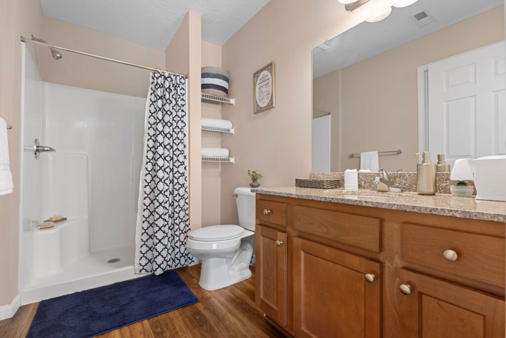 Bathroom with a walk-in shower, patterned curtain, toilet, wooden vanity with countertop, wall shelves, and mirror. Blue and white accents and neatly arranged toiletries are visible.