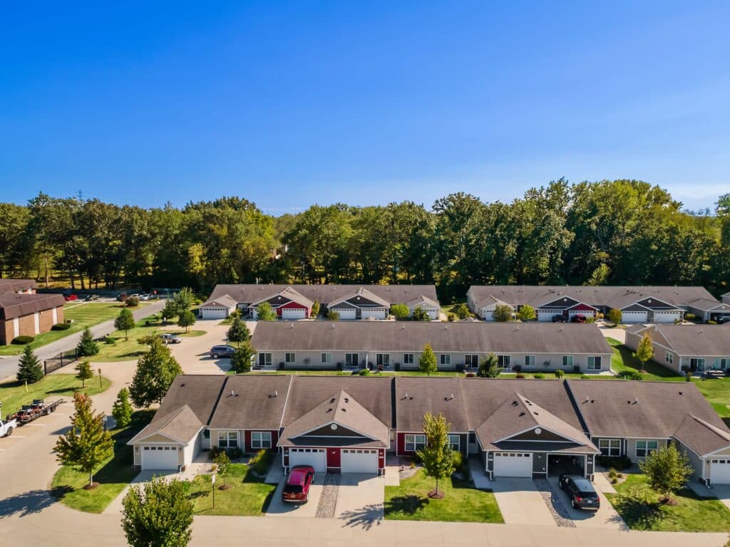 Aerial view of a suburban neighborhood with rows of single-story homes, driveways, and parked cars, bordered by trees under a clear blue sky.