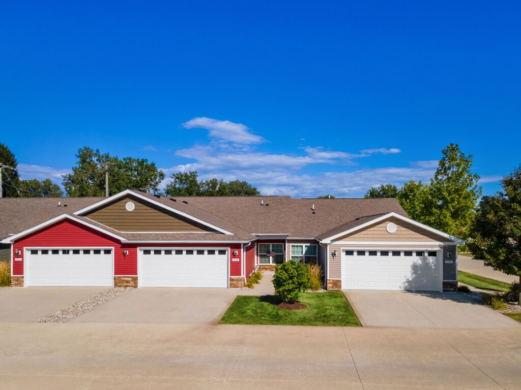 A row of single-story townhouses with attached double garages, red and beige siding, and a clear blue sky overhead.