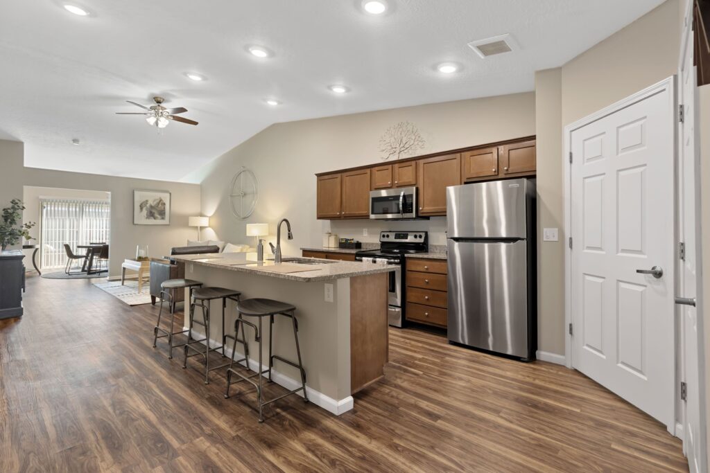 Open-concept kitchen with stainless steel appliances, brown cabinets, an island with stools, and wood flooring, leading to a dining and living area with natural light.
