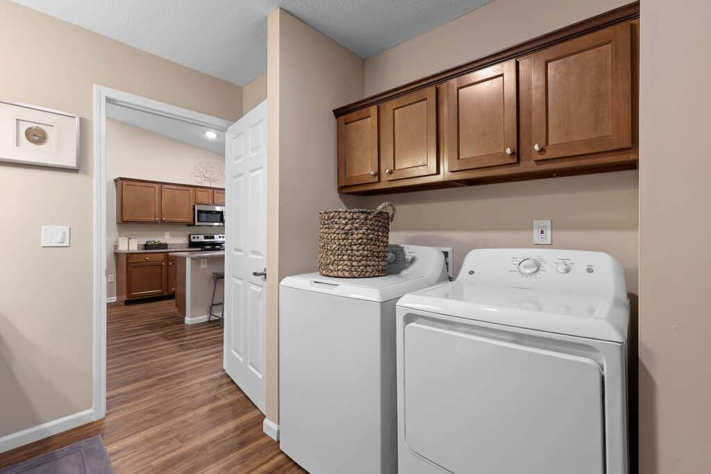 Laundry room with a washing machine, dryer, basket, and wooden cabinets, adjacent to a kitchen with similar wood cabinetry and hardwood floors.