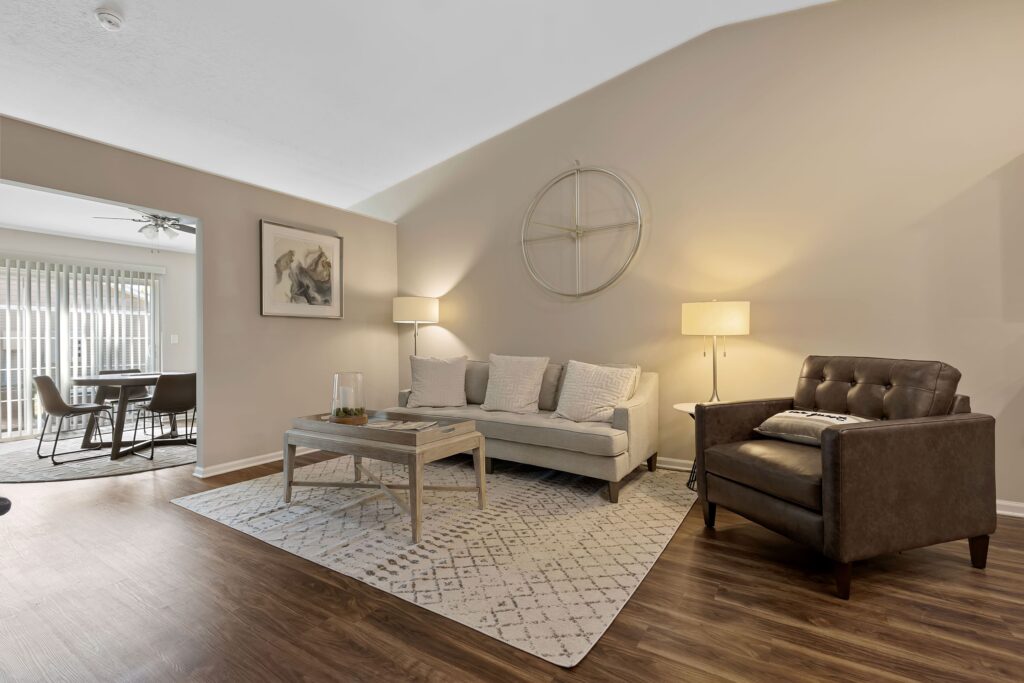A modern living room with a gray sofa, brown leather armchair, two lamps, a coffee table, and wall art. A dining area is visible through an open doorway.