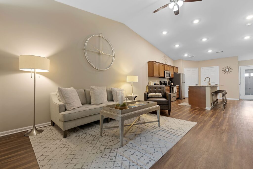 Open-concept living room and kitchen with neutral tones, featuring a sofa, armchair, coffee table, floor lamp, and an island with wooden cabinets and stainless steel appliances.