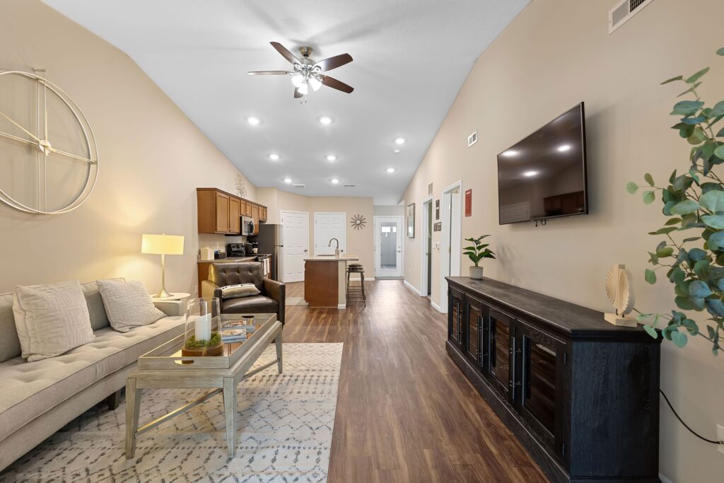 Modern open-concept living room and kitchen with wood flooring, neutral furnishings, wall-mounted TV, and ceiling fan with recessed lighting.