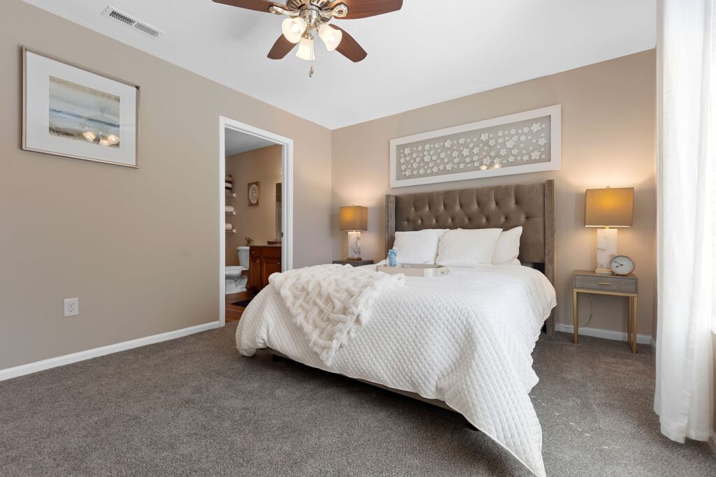 A neatly made bed with white bedding in a carpeted bedroom, featuring two bedside tables with lamps and neutral-colored walls. A ceiling fan and framed art are visible.