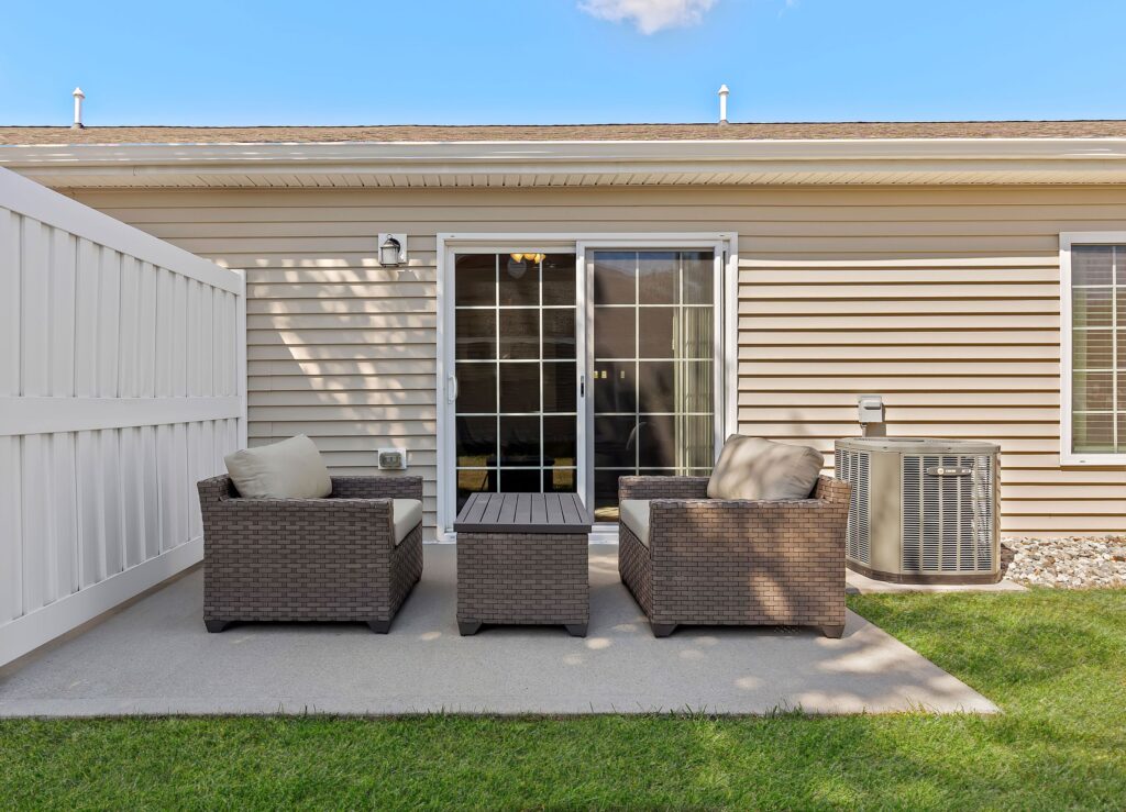 Two wicker chairs and a matching table sit on a small concrete patio outside a beige house with sliding glass doors and an air conditioning unit.