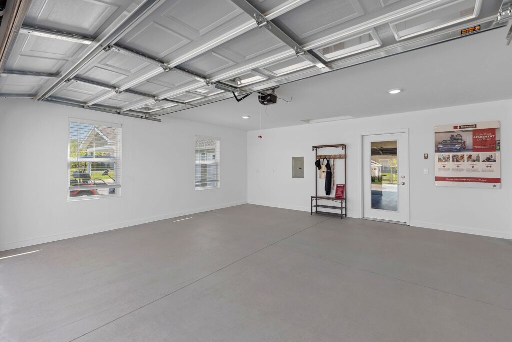 A clean, spacious two-car garage with gray flooring, white walls, three windows, a door to the outside, and a wall-mounted coat rack with a bench.