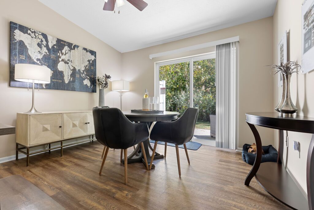 A dining area with a round table and four chairs, wood flooring, a sideboard, wall art, large window with vertical blinds, and a view of greenery outside.