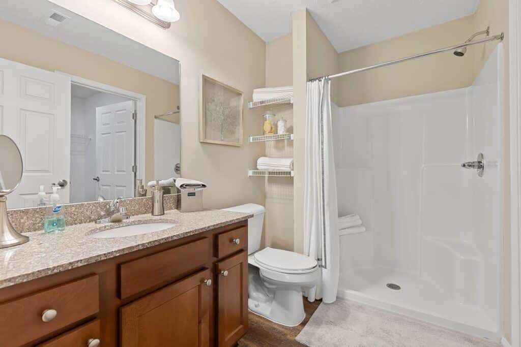 Bathroom with wooden vanity, granite countertop, single sink, toilet, walk-in shower with white curtain, wall shelves with towels, and neutral beige walls.