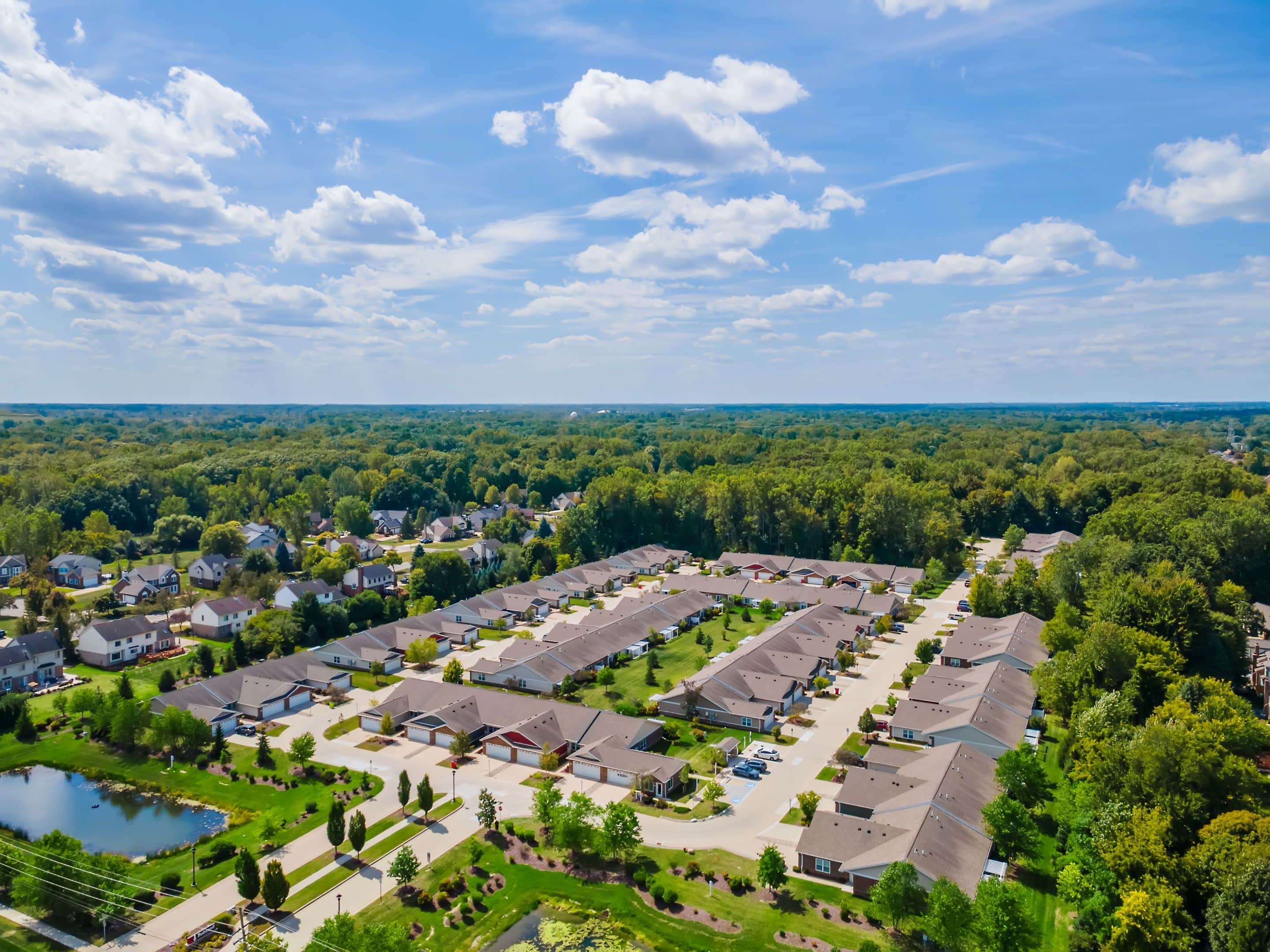 Aerial view of a suburban neighborhood with rows of single-family homes, green lawns, a pond, and surrounding trees under a partly cloudy sky.