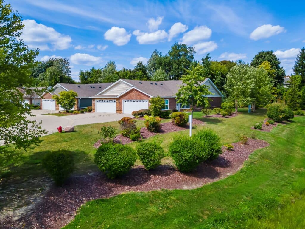 Single-story brick duplex residences with attached garages, surrounded by landscaped lawns, shrubs, and trees under a blue sky with scattered clouds.