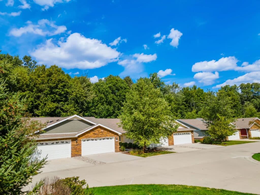 Row of suburban single-story houses with attached garages and trees in the background under a clear blue sky with scattered clouds.