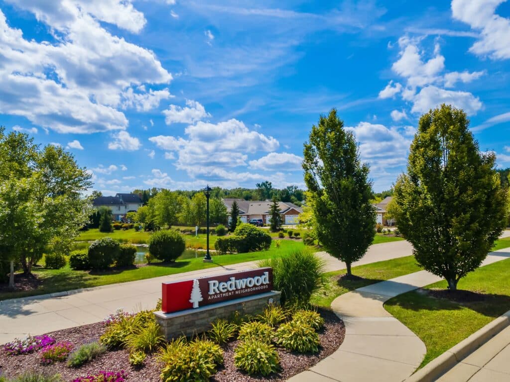 A landscaped entrance sign for Redwood Apartment Neighborhoods stands beside a sidewalk, with trees, grass, and residential buildings in the background under a blue, partly cloudy sky.