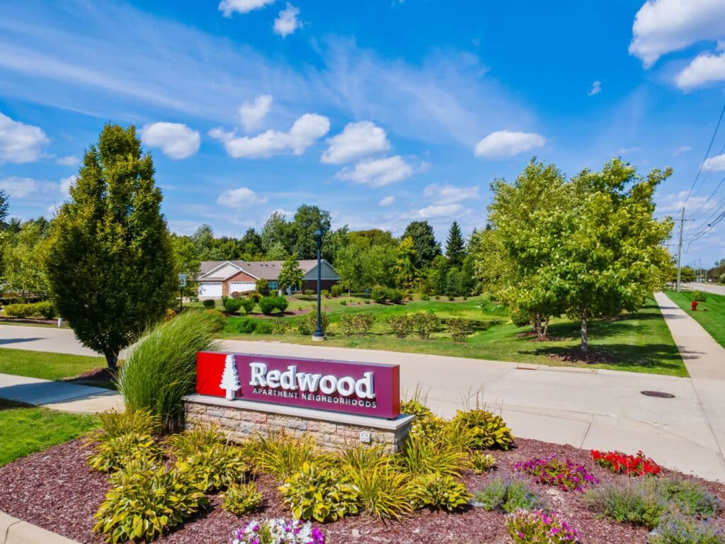 A landscaped entrance with a red sign reading "Redwood Apartment Neighborhoods," surrounded by greenery and flowers on a sunny day.