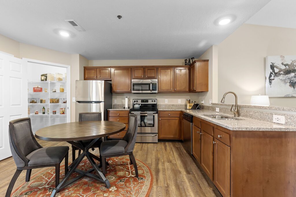 Modern kitchen with stainless steel appliances, wooden cabinets, granite countertops, round dining table with four chairs, and an open pantry stocked with food.