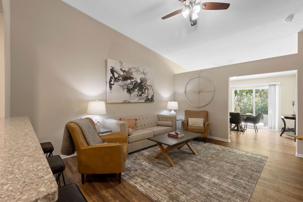 A modern living room with a beige sofa, two armchairs, a coffee table, abstract wall art, a large wall clock, and hardwood floors, leading to a dining area with glass doors.