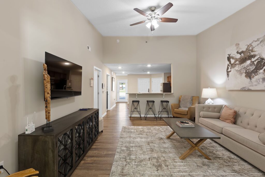 Modern living room with neutral tones, sofa, coffee table, TV on a console, abstract wall art, and a view of kitchen with bar stools in the background. Ceiling fan and recessed lighting overhead.