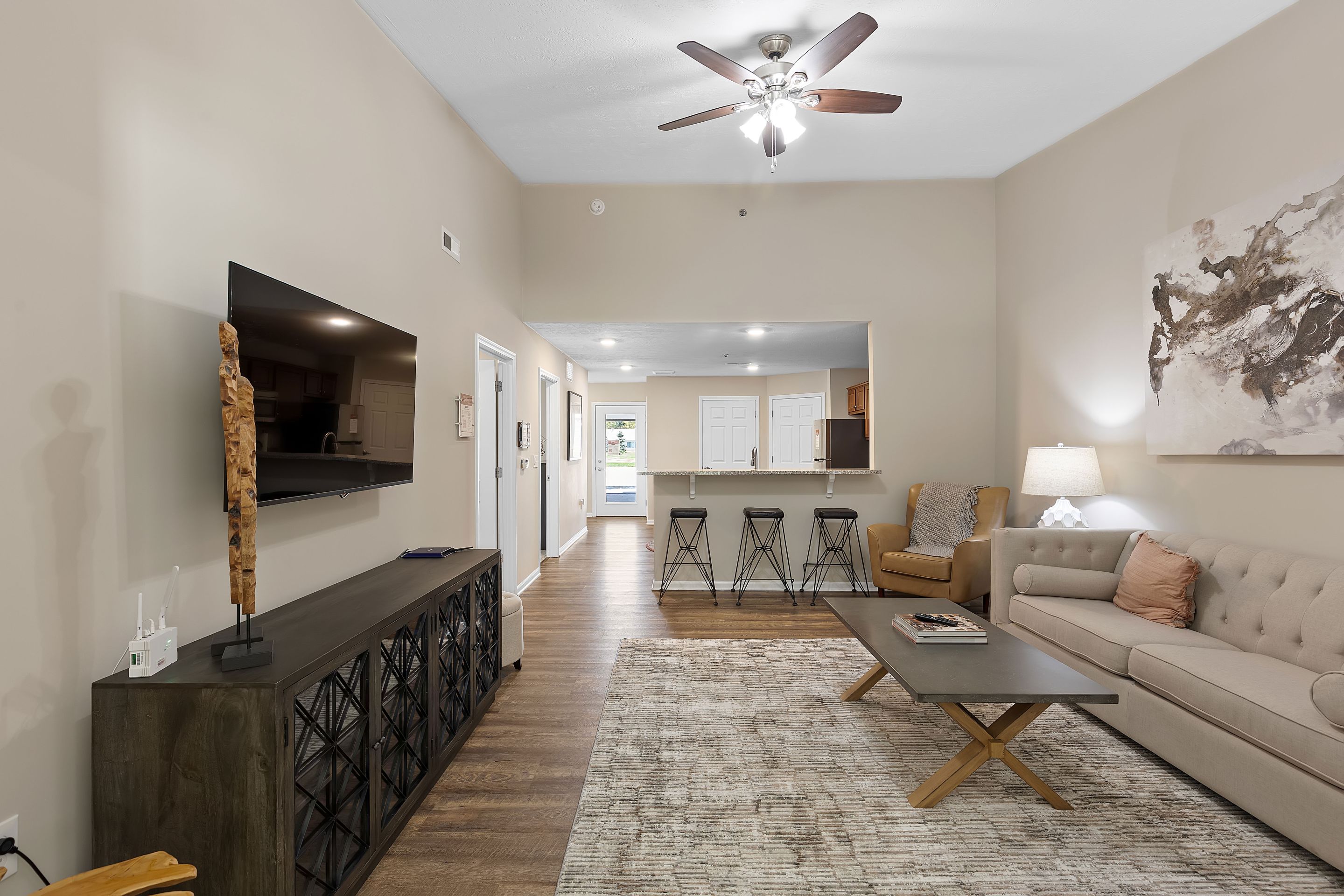 Modern living room with neutral tones, sofa, coffee table, TV on a console, abstract wall art, and a view of kitchen with bar stools in the background. Ceiling fan and recessed lighting overhead.