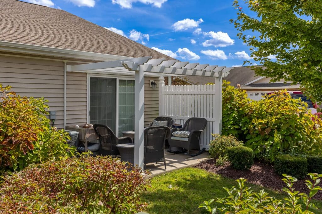 A small patio with a white pergola, four black chairs, and a table, surrounded by shrubs and greenery next to a beige house.